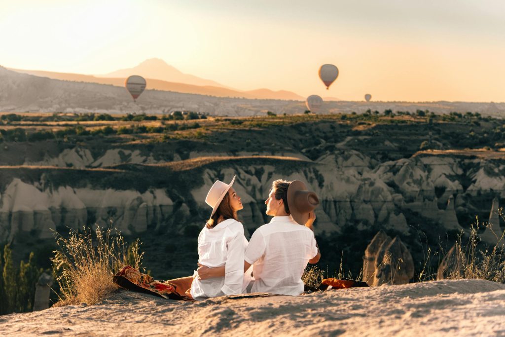 A couple sits in Cappadocia, watching hot air balloons at sunset.