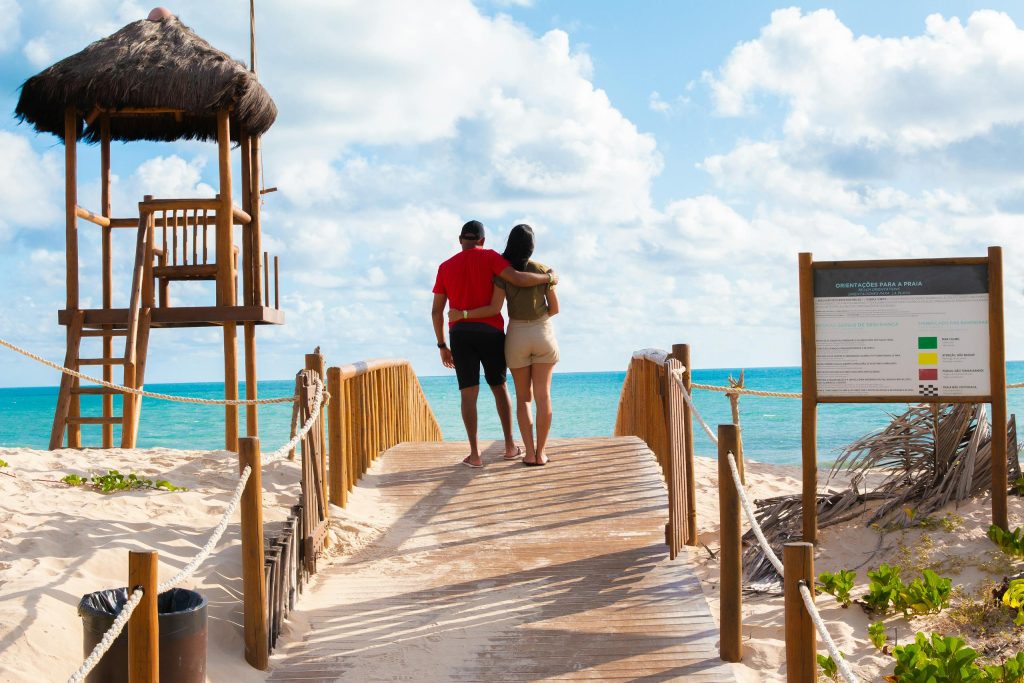 A couple walks hand in hand on a footbridge towards a sunny beach in Natal, Brazil, with clear blue skies.