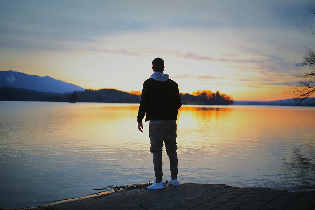 A man stands by a tranquil lake at sunset, reflecting the serene sky.