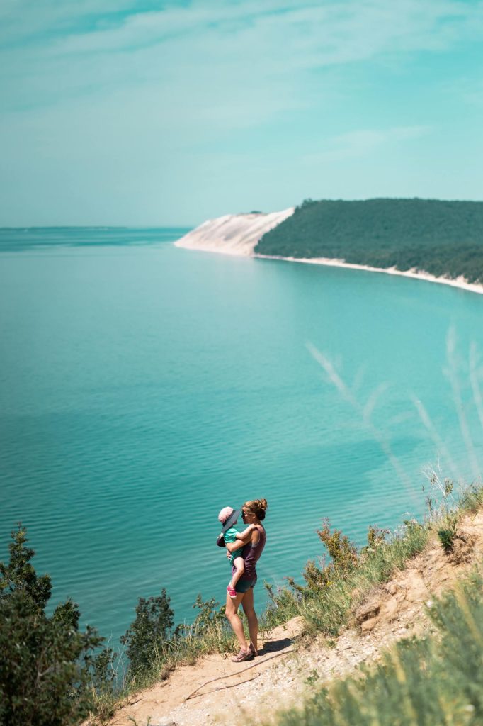 A mother holds her child overlooking tranquil turquoise waters and scenic dunes on a sunny day.