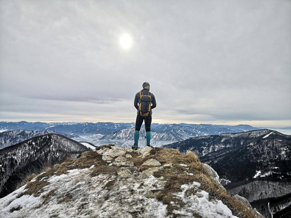 A solitary hiker with a backpack stands on a snowy mountain summit, gazing at the vast winter landscape.