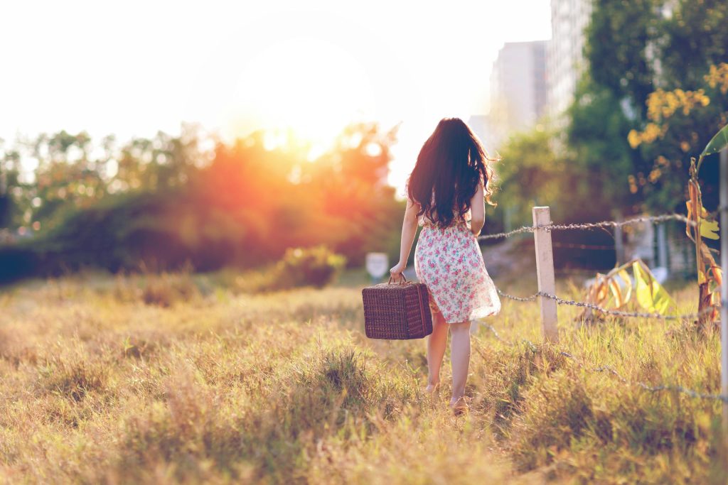 A woman walks through a grassy field with a basket during sunset, creating a serene and warm atmosphere.