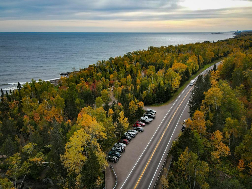Breathtaking aerial view of a forested lakeshore during fall with vibrant foliage and parked cars along the road.