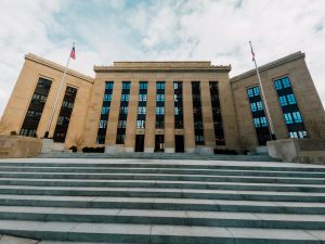 Federal style government building with flags and stairway under cloudy sky.