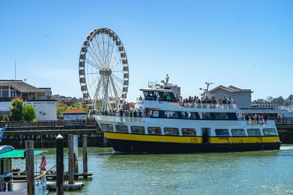 Ferry filled with passengers sailing past a Ferris wheel on a sunny day in San Francisco Bay.