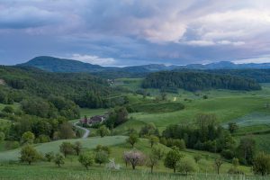 Lush green countryside with rolling hills under a dramatic cloudy sky during summer.