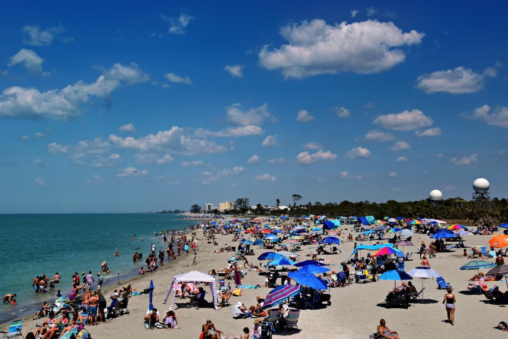 People enjoying a sunny day at a crowded beach in Venice, Florida, under colorful umbrellas.
