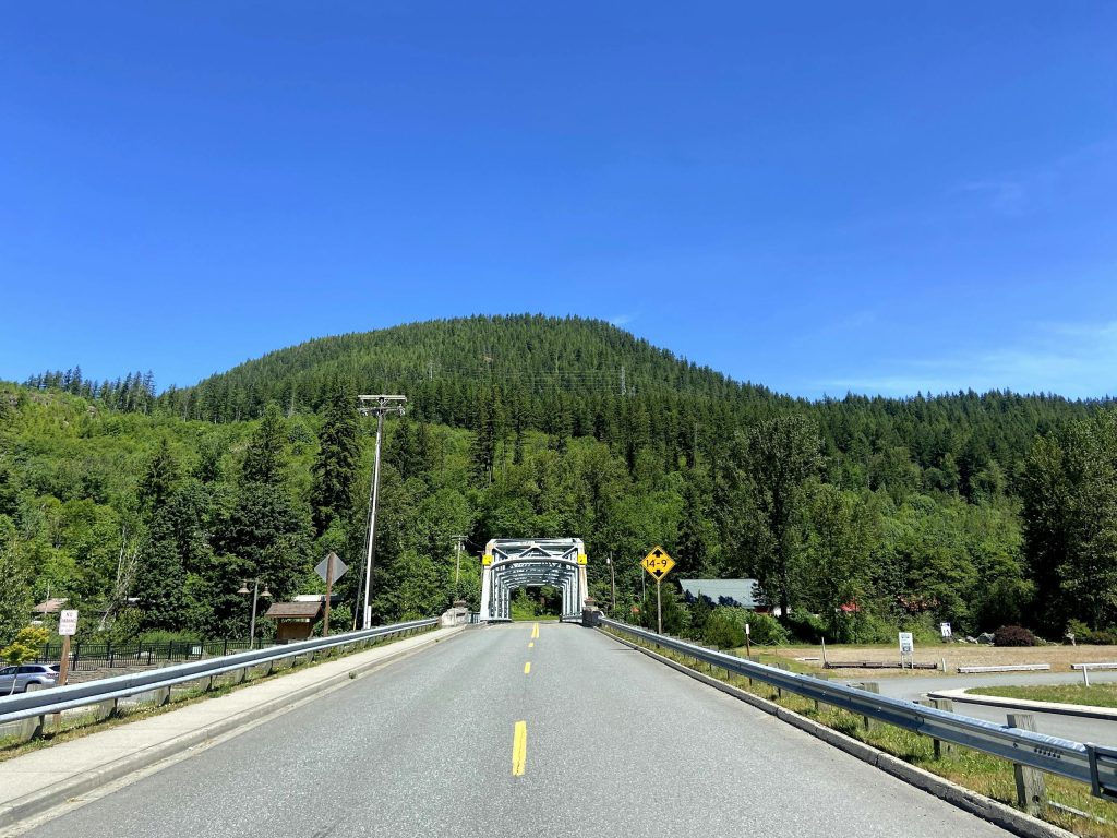 View of a scenic road leading to a bridge, surrounded by lush green forests and clear blue sky.