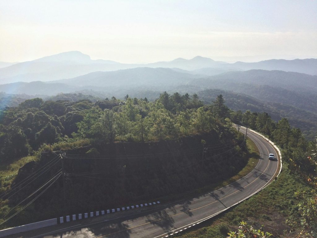 Aerial view of a winding road through lush green forest with misty mountain backdrop in Chiang Mai, Thailand.