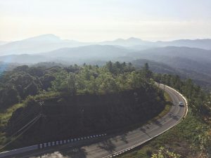 Aerial view of a winding road through lush green forest with misty mountain backdrop in Chiang Mai, Thailand.