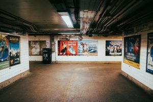 Empty subway station with vibrant posters on white tiled walls and overhead pipes visible.