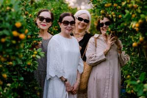 Four senior women smiling and enjoying a day in a lush green citrus orchard, wearing sunglasses.