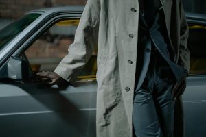 Mysterious figure in trench coat leans against a vintage car, evoking a noir detective vibe.