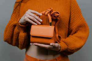 Woman holding an orange leather mini handbag against an orange sweater in a studio setting.
