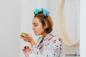 Woman with hair curlers applies skincare cream in front of a bathroom mirror.