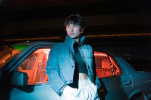 Young woman leaning against a car with colorful lighting creating a dramatic night scene.